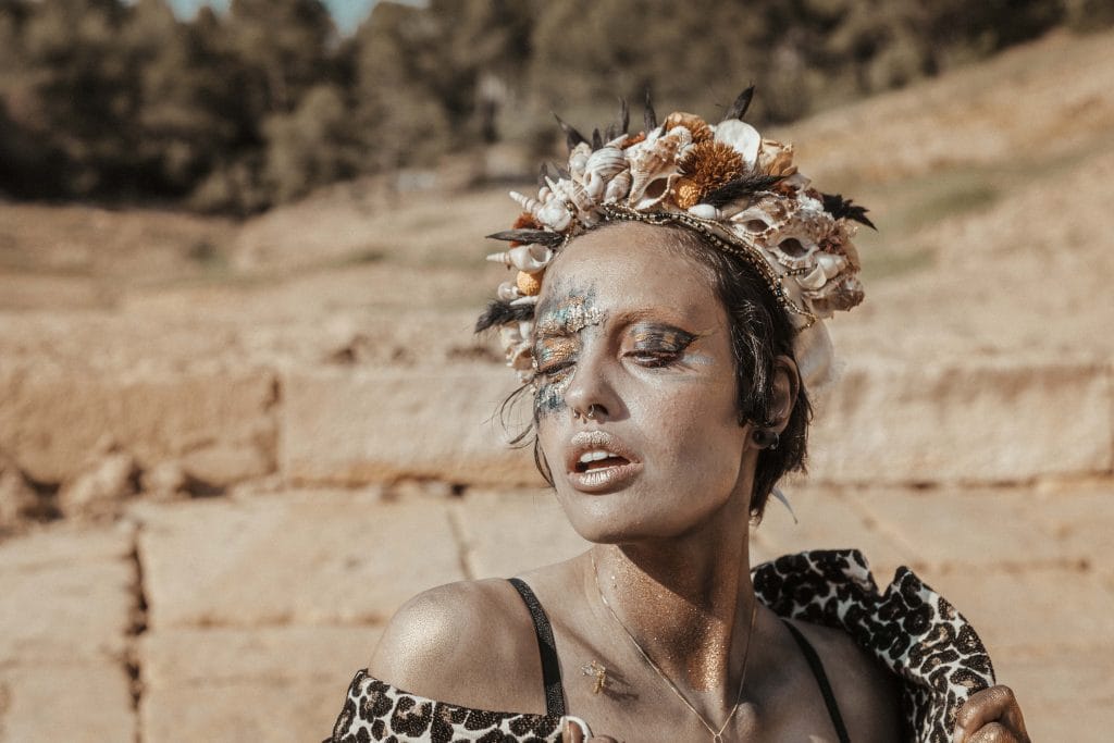Une personne se tient à l'extérieur, les yeux fermés, ornée d'un coquillage aux couleurs vives et d'un couvre-chef en plumes. Son visage est orné d'un maquillage bleu et orange saisissant, parfait pour un portrait photographique unique. Elle porte un vêtement à imprimé léopard sur fond d'arbres et de terrain rocheux.