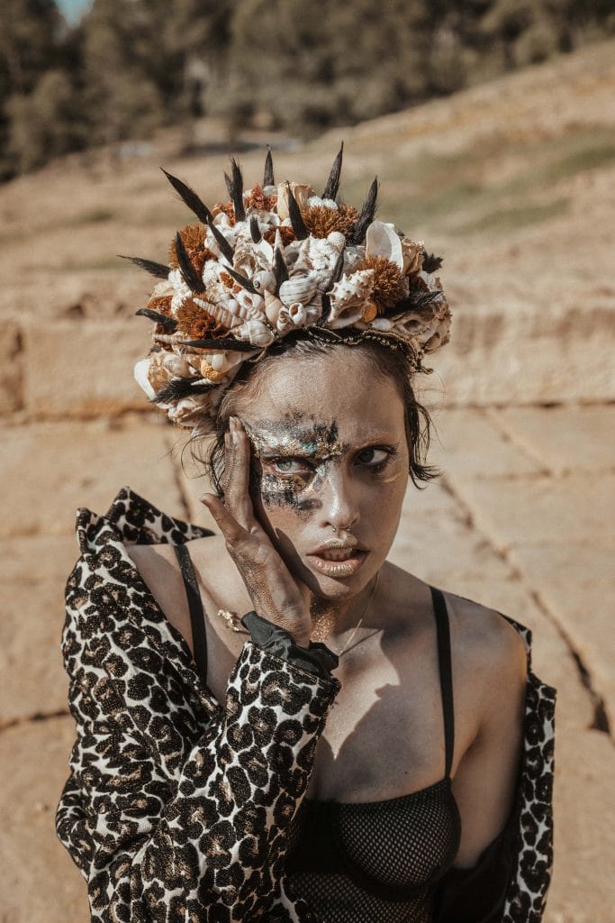 Une personne portant un manteau à imprimé léopard et une coiffe en coquillages et plumes pose avec éloquence à l'extérieur par une journée ensoleillée, incarnant l'essence vibrante de la Guadeloupe. Son visage est orné d'un maquillage artistique, avec des formes sombres autour des yeux et du front, tandis qu'une main touche gracieusement son visage.