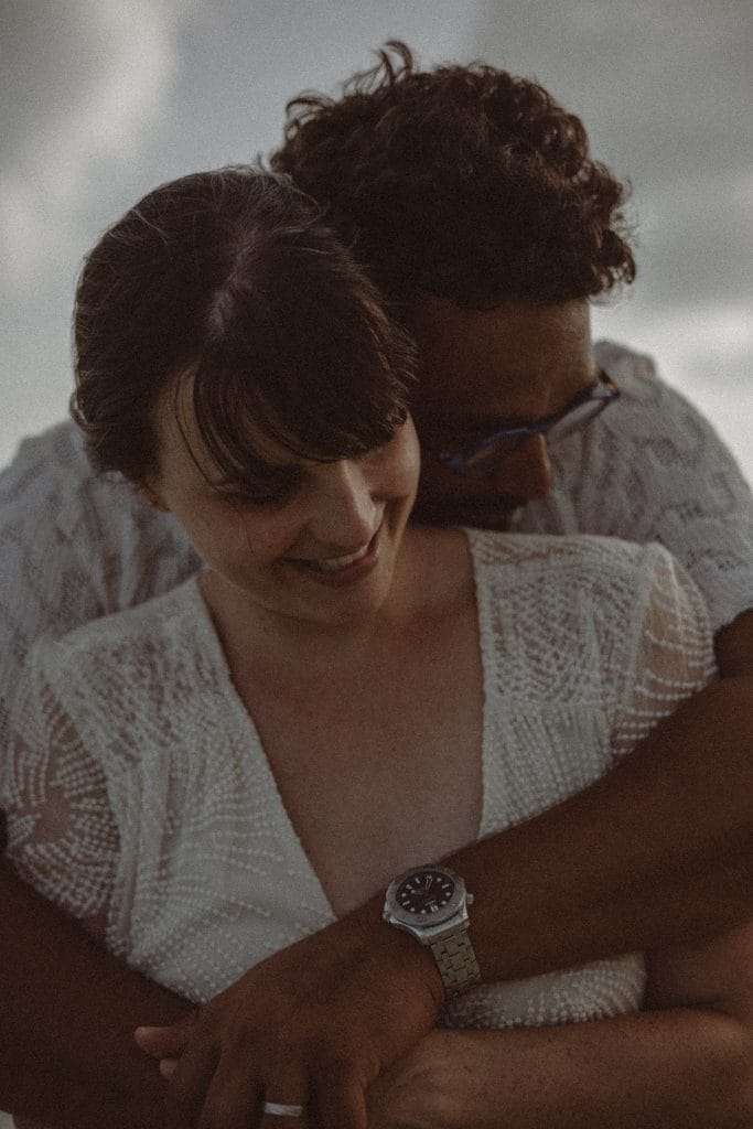 Un couple s'embrasse chaleureusement, l'homme enlaçant la femme par derrière. Tous deux portent des vêtements blancs à motifs et sourient légèrement. Avec sa montre-bracelet et ses lunettes de soleil, il semble prêt à être photographié pour un mariage en Guadeloupe. La scène doucement éclairée suggère un moment intime et paisible.
