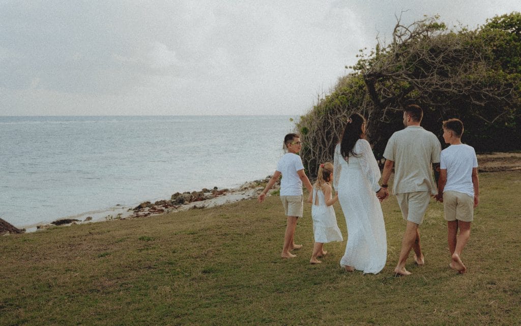 Famille de cinq personnes en tenue blanche et beige marchant pieds nus sur l'herbe face à la mer en Guadeloupe, photo CW Photographe